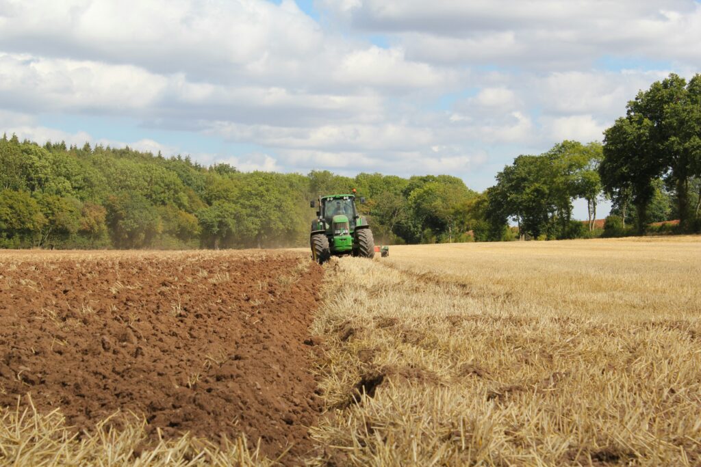 A green tractor plowing a field by a woodland.